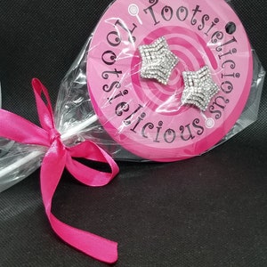 May include: A pair of silver star-shaped earrings with clear rhinestones, packaged in a pink circle with the text "Tootsielicious" on it.