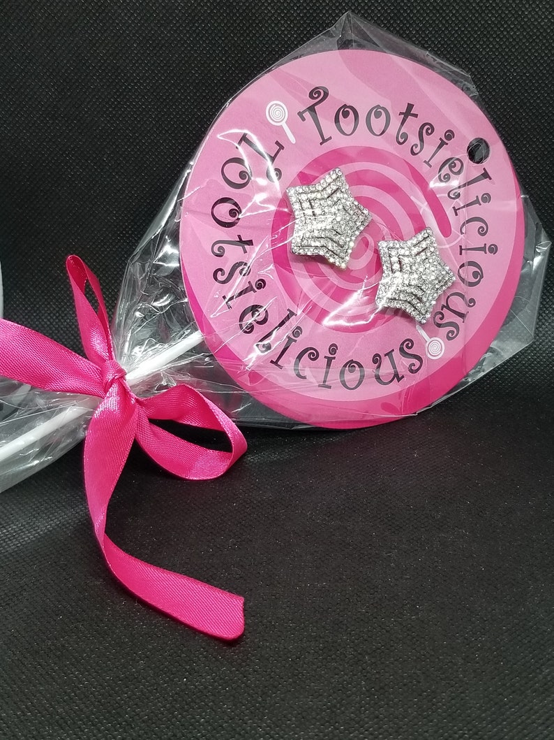 May include: A pair of silver star-shaped earrings with clear rhinestones, packaged in a pink circle with the text "Tootsielicious" on it.