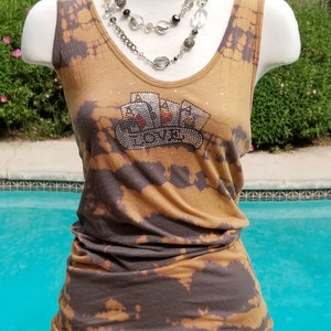 May include: A brown and black tie-dye tank top with a rhinestone design of playing cards and the word "Ace" on the front. The tank top is being modeled on a white mannequin with a layered necklace of clear and black beads.