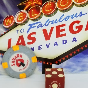 May include: A red and white dice with the word "Sahara" and "Las Vegas" printed on it. The dice is sitting on a white surface in front of a vintage postcard with a neon sign that reads "Welcome to Fabulous Las Vegas, Nevada".