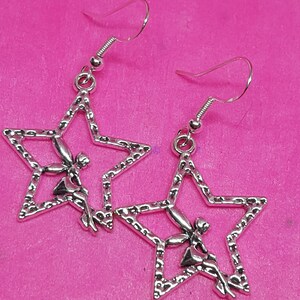 May include: A pair of silver earrings featuring a star design with a fairy sitting inside. The earrings have a hook closure.
