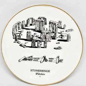 May include: White ceramic plate with a gold rim featuring a black and white illustration of Stonehenge. The text "STONEHENGE Wiltshire" is printed below the illustration. A decorative item.