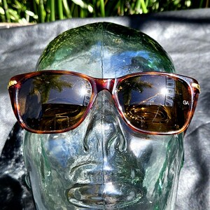 May include: Tortoiseshell-patterned sunglasses with dark brown lenses and gold-tone metal accents at the hinges. The right lens features the letters GA printed in white. The frames have a rectangular shape with rounded edges and a bridge.