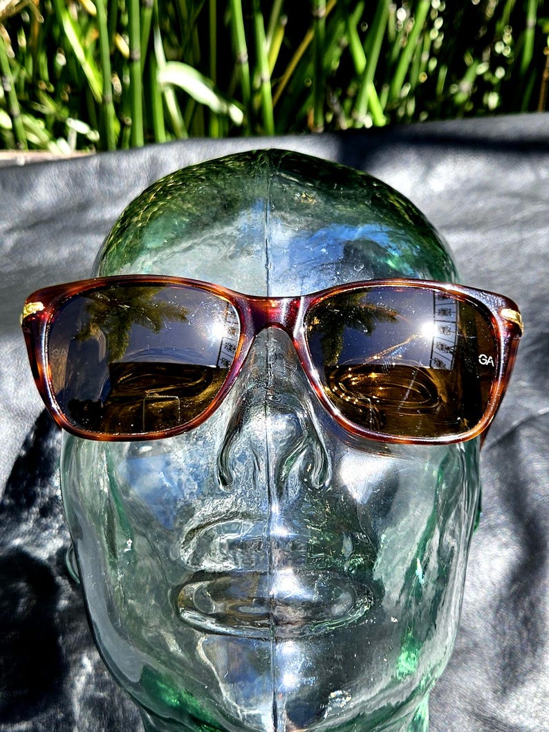 May include: Tortoiseshell-patterned sunglasses with dark brown lenses and gold-tone metal accents at the hinges. The right lens features the letters GA printed in white. The frames have a rectangular shape with rounded edges and a bridge.