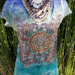 May include: A turquoise blue, white, and purple short-sleeved shirt with a colorful rhinestone design. The shirt has a V-neck and a distressed look.
