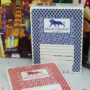 May include: Two decks of playing cards with a blue and red background. Both decks feature a lion logo with the text "MGM GRAND. LAS VEGAS". The blue deck has a barcode and the text "GAME/TABLE#" and "DATE". The red deck has a barcode and the text "Deck of Cards Was Used in Actual Play in MGM".