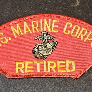 May include: A red patch with gold embroidered text that reads "U.S. MARINE CORPS RETIRED" and a gold embroidered emblem of the Marine Corps.