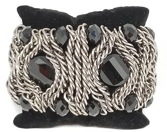 May include: A wide bracelet featuring a woven silver-tone metal design, interspersed with faceted black beads. The bracelet has a bold, chunky appearance, with a central section of twisted metal. The black beads add contrast and visual interest.
