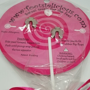 May include: A pink and white lollipop with a silver charm attached. The charm is shaped like a foot with two prongs. The lollipop is wrapped in clear plastic and tied with a pink ribbon. The packaging says "www.Tootsielicious.com" and "Sweet treats for the feet".