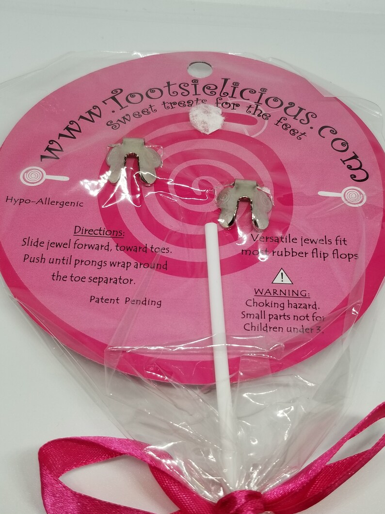 May include: A pink and white lollipop with a silver charm attached. The charm is shaped like a foot with two prongs. The lollipop is wrapped in clear plastic and tied with a pink ribbon. The packaging says "www.Tootsielicious.com" and "Sweet treats for the feet".