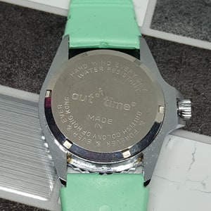 May include: A silver-toned wristwatch with a green band. The watch face is not visible. The back of the watch has the text "Out of time&reg;" and "Made in British Colony of Hong Kong & Forever & Ever &" printed on it.