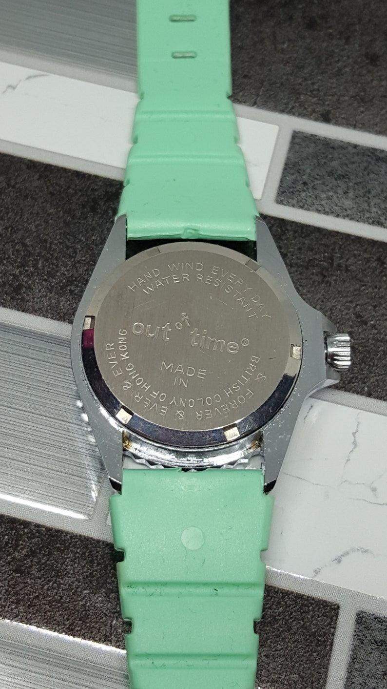 May include: A silver-toned wristwatch with a green band. The watch face is not visible. The back of the watch has the text "Out of time&reg;" and "Made in British Colony of Hong Kong & Forever & Ever &" printed on it.