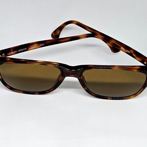 May include: Tortoiseshell-patterned Giorgio Armani sunglasses with brown lenses. The inner temple arm features the text Giorgio Armani, Made in Italy, and the model numbers 810 050 140. The frames have a classic rectangular shape with gold-tone accents.