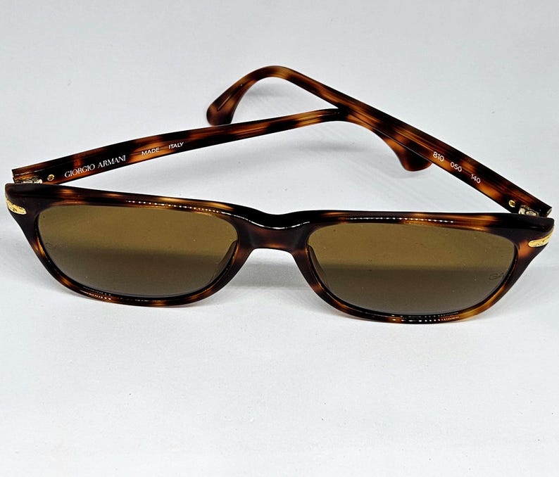 May include: Tortoiseshell-patterned Giorgio Armani sunglasses with brown lenses. The inner temple arm features the text Giorgio Armani, Made in Italy, and the model numbers 810 050 140. The frames have a classic rectangular shape with gold-tone accents.