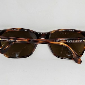 May include: Tortoiseshell-patterned sunglasses with brown lenses and gold-tone metal accents on the hinges. The temples are inscribed with the text GIORGIO ARMANI in gold lettering. The frames feature a classic rounded rectangular shape.