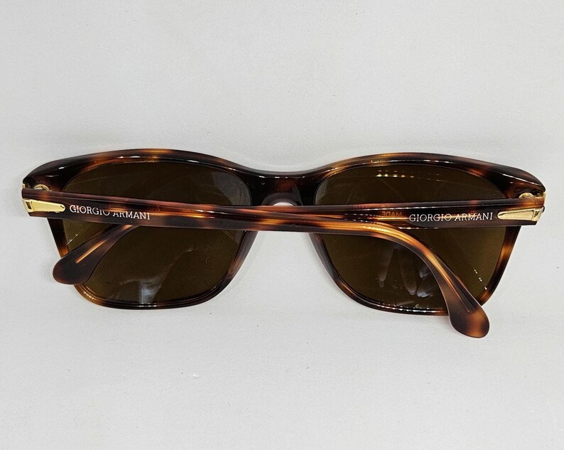 May include: Tortoiseshell-patterned sunglasses with brown lenses and gold-tone metal accents on the hinges. The temples are inscribed with the text GIORGIO ARMANI in gold lettering. The frames feature a classic rounded rectangular shape.