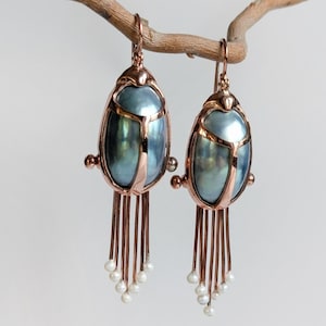 Pode incluir: Um par de brincos cor de cobre com um design de escaravelho. Os brincos apresentam um centro azul-esverdeado perolado, detalhes cor de cobre e fios pendurados com pequenas pérolas brancas. Os brincos estão pendurados em um galho marrom.