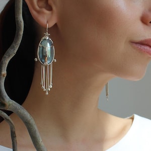 Pode incluir: Brincos prateados com uma pedra central ovalada e iridescente e fios pendurados. Os brincos são usados por uma pessoa com uma blusa branca. Uma peça de joalharia única.
