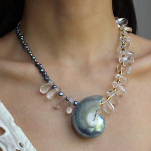 May include: A handcrafted necklace featuring a large, iridescent, blue-gray pearl pendant. The necklace incorporates clear, faceted crystal beads and smaller, dark gray beads, strung on a delicate chain. The design is elegant and eye-catching.