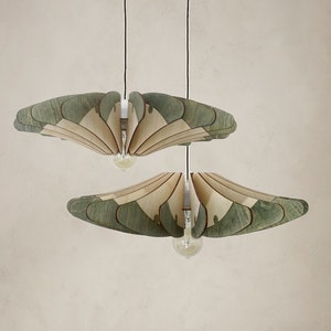 May include: Two wooden pendant lights with a layered, fan-like design in shades of green and beige hang from the ceiling above a brown wooden chair and table. The chair has a woven cane seat and back and is covered with a gray throw blanket.