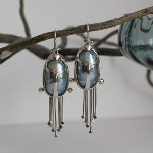 Pode incluir: Um par de brincos prateados com um design único. Cada brinco apresenta uma grande pedra oval azul perolada emoldurada em prata. Delicados fios de prata pendem da parte inferior, adornados com pequenas contas coloridas. Os brincos são suspensos por um gancho simples.