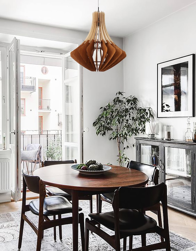 May include: A wooden pendant light fixture with a layered, geometric design hangs above a round wooden dining table. The room features a rug, chairs, and a cabinet. A framed picture and a plant are also visible.