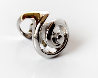 Könnte beinhalten: Ein polierter Silberring mit einem einzigartigen, wirbelnden Design. Der Ring hat an einem Ende eine Spiralform und ein gebogenes Band. Die reflektierende Oberfläche des Rings fängt das Licht ein und hebt seine glatte, metallische Textur hervor. Schmuck.