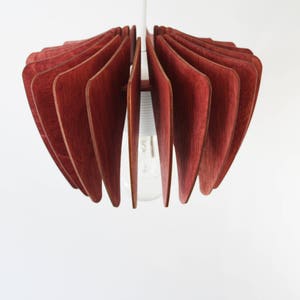 May include: A red wooden pendant light fixture with a unique, geometric design. The light fixture is made of thin, curved pieces of wood that are arranged in a fan shape. The light fixture is suspended from the ceiling by a single cord.