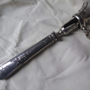 Puede incluir: Un abridor de botellas de vino plateado vintage. El abridor presenta un mango largo y rectangular con detalles florales y de volutas. La parte superior tiene un mecanismo con brazos curvos. El mango tiene un diseño grabado. El abridor está sobre un fondo blanco.