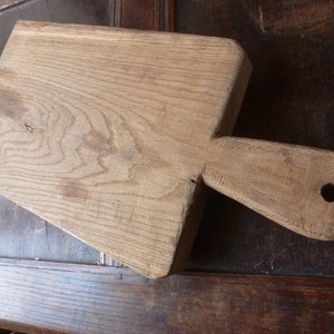 Op de afbeelding: Een groot, rustiek houten snijplank met een handvat en een gat. De plank is gemaakt van lichtbruin hout en heeft een natuurlijke, verweerde afwerking.