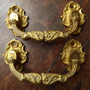 Pode incluir: Duas puxadores de gaveta de metal dourado ornamentado com um design floral decorativo. As puxadores têm uma forma curva e são projetadas para serem montadas em um armário ou gaveta.