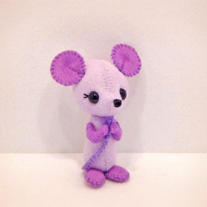 May include: A purple felt mouse toy with black eyes and a purple bow tie. The mouse is standing on a white surface.