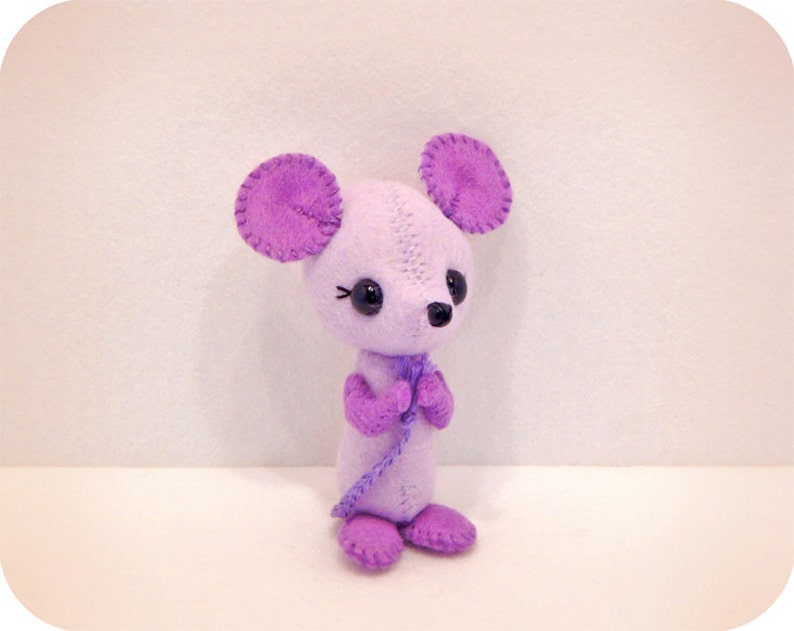 May include: A purple felt mouse toy with black eyes and a purple bow tie. The mouse is standing on a white surface.