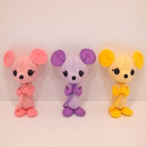 May include: Three plush mouse toys, each in a different colour: pink, purple and yellow. The toys have black eyes and are standing upright with their arms crossed.