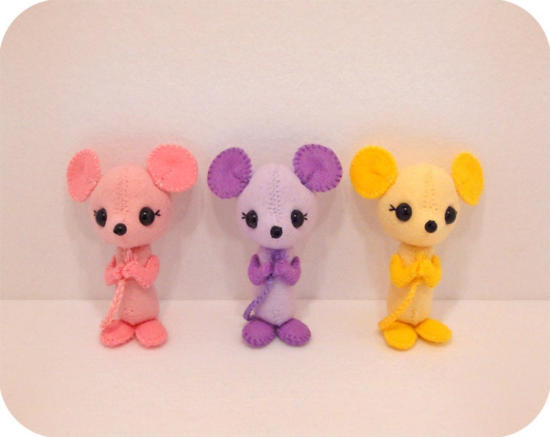 May include: Three plush mouse toys, each in a different colour: pink, purple and yellow. The toys have black eyes and are standing upright with their arms crossed.