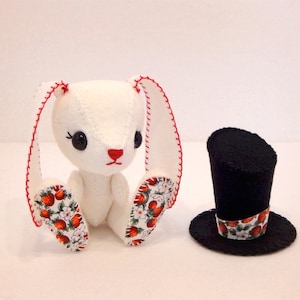 May include: A white felt bunny with red stitching and strawberry patterned fabric on its ears and feet. The bunny is wearing a black felt top hat with a white and red strawberry patterned band.