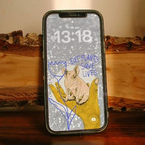 Könnte beinhalten: Ein Smartphone mit einem Sperrbildschirm, der eine digitale Illustration eines Schweins auf einer gelben Hand, eine blaue Strichzeichnung einer Blume und den Text EAT PLANTS SAVE LIVES zeigt. Das Display zeigt Datum Fri 26 Jun und Uhrzeit 13:18.