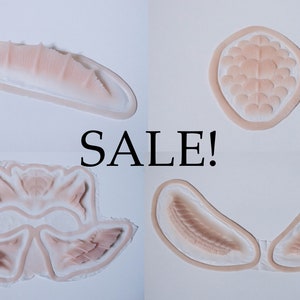 May include: Four prosthetic makeup pieces in various shapes and sizes, with a flesh-toned colour and a white border. The word "SALE!" is displayed in black, bold text.