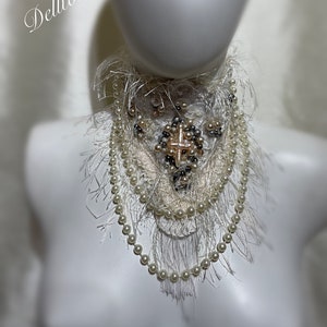 May include: A white, fluffy, beaded and fringed choker necklace with a cross-shaped embellishment. The necklace is on a white mannequin bust. The text "Dellton Couture" is visible in the background.