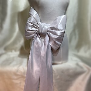 May include: A large white satin bow with long tails. The bow is tied around a white mannequin torso. The text "Dellton Couture" is visible in the top left corner of the image.