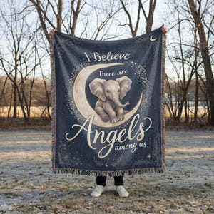 May include: A dark blue woven throw blanket featuring a central illustration of a baby elephant sitting on a crescent moon. The text reads I Believe There are Angels among us in a decorative script font, surrounded by celestial and floral motifs.