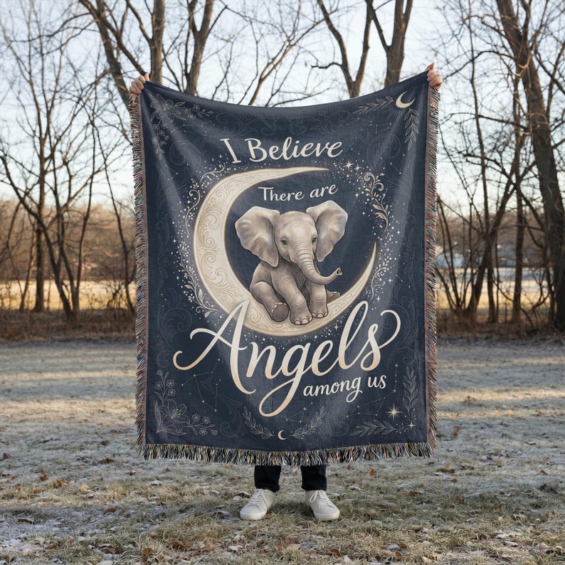 May include: A dark blue woven throw blanket featuring a central illustration of a baby elephant sitting on a crescent moon. The text reads I Believe There are Angels among us in a decorative script font, surrounded by celestial and floral motifs.