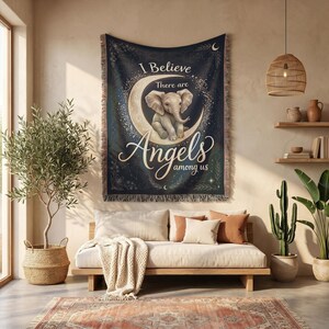 May include: A rectangular woven throw blanket featuring a dark blue celestial background with a crescent moon and a central baby elephant illustration. The text reads I Believe There are Angels among us in white and gold script lettering.