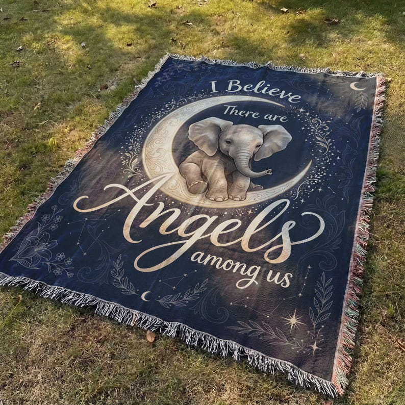 May include: A rectangular woven throw blanket featuring a dark blue background with a central illustration of a baby elephant sitting on a crescent moon. The text reads I Believe There are Angels among us. The edges have a decorative fringe.