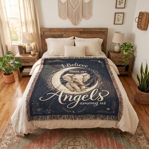 May include: A dark blue woven throw blanket featuring a central graphic of a baby elephant sitting on a crescent moon. The text reads I Believe There are Angels among us. The blanket has a fringed edge and decorative floral and star accents.