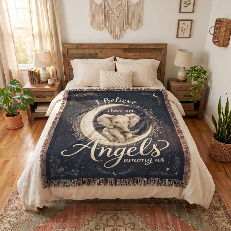 May include: A dark blue woven throw blanket featuring a central graphic of a baby elephant sitting on a crescent moon. The text reads I Believe There are Angels among us. The blanket has a fringed edge and decorative floral and star accents.