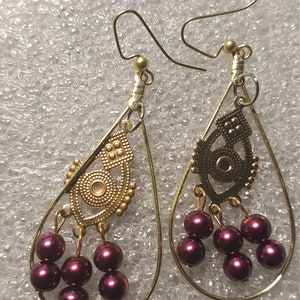 May include: A pair of gold dangle earrings with a burgundy pearl drop. The earrings feature a gold filigree design and are suspended from a gold hoop.
