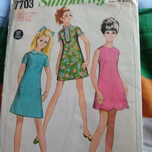 May include: A vintage Simplicity 7703 sewing pattern envelope featuring illustrations of three dresses. One is a solid teal shift dress with pockets, one is a green floral print dress with a ruffled collar, and one is a solid pink scalloped hem dress.