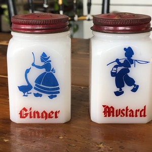May include: Two white ceramic salt and pepper shakers with red lids. The shakers have blue and white illustrations of people in traditional Dutch clothing. The shaker on the left says "Ginger" and the shaker on the right says "Mustard".