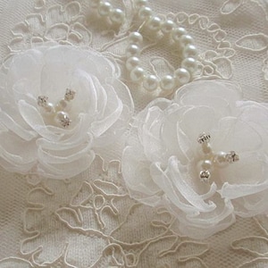 May include: Two white fabric flowers with pearl and rhinestone embellishments on a white lace background.